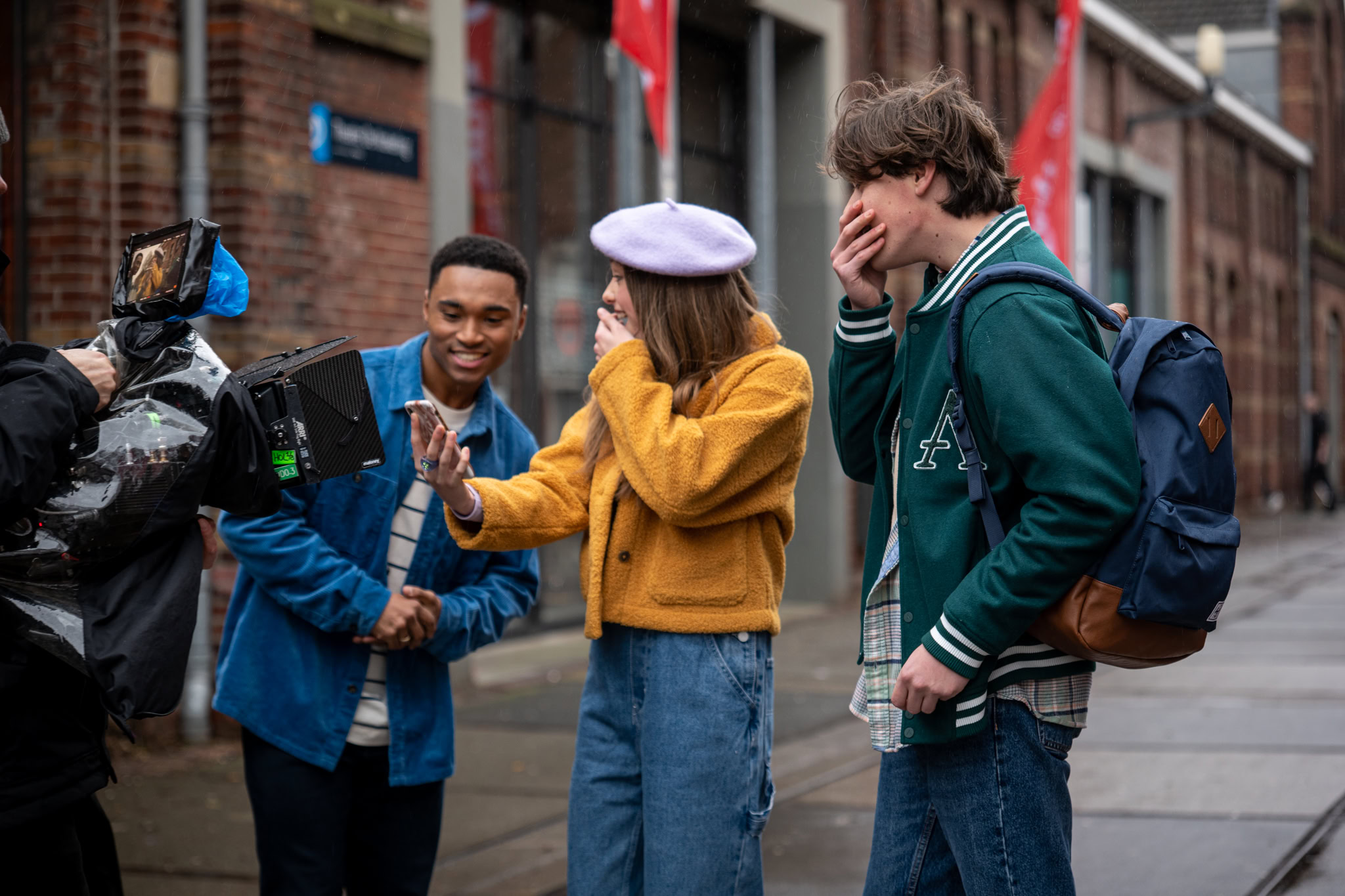Behind-the-scenes of an on-location film shoot — a camera operator with a rain-covered cinema camera films three young actors on a wet city street